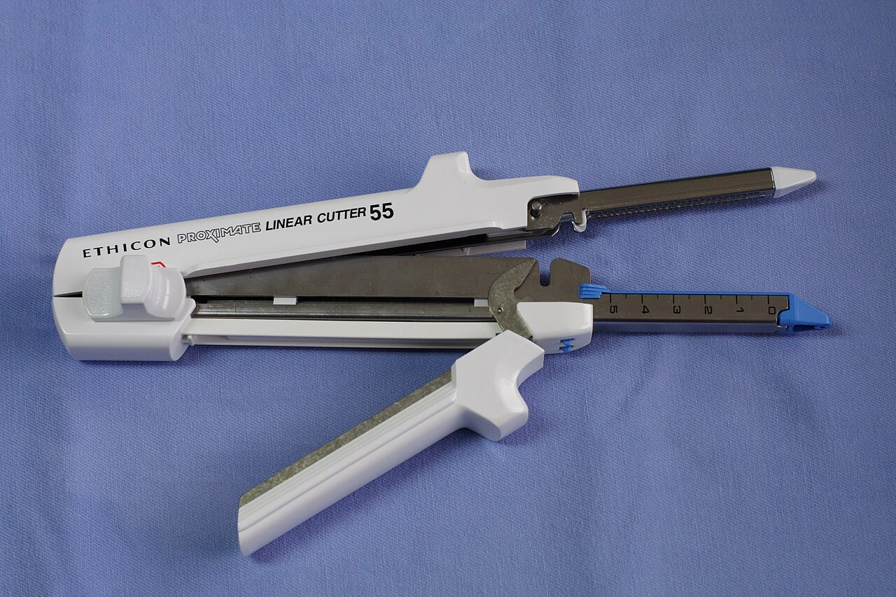A long pale handled device has parallel distal jaws, with a separate elongated cartridge beside it.