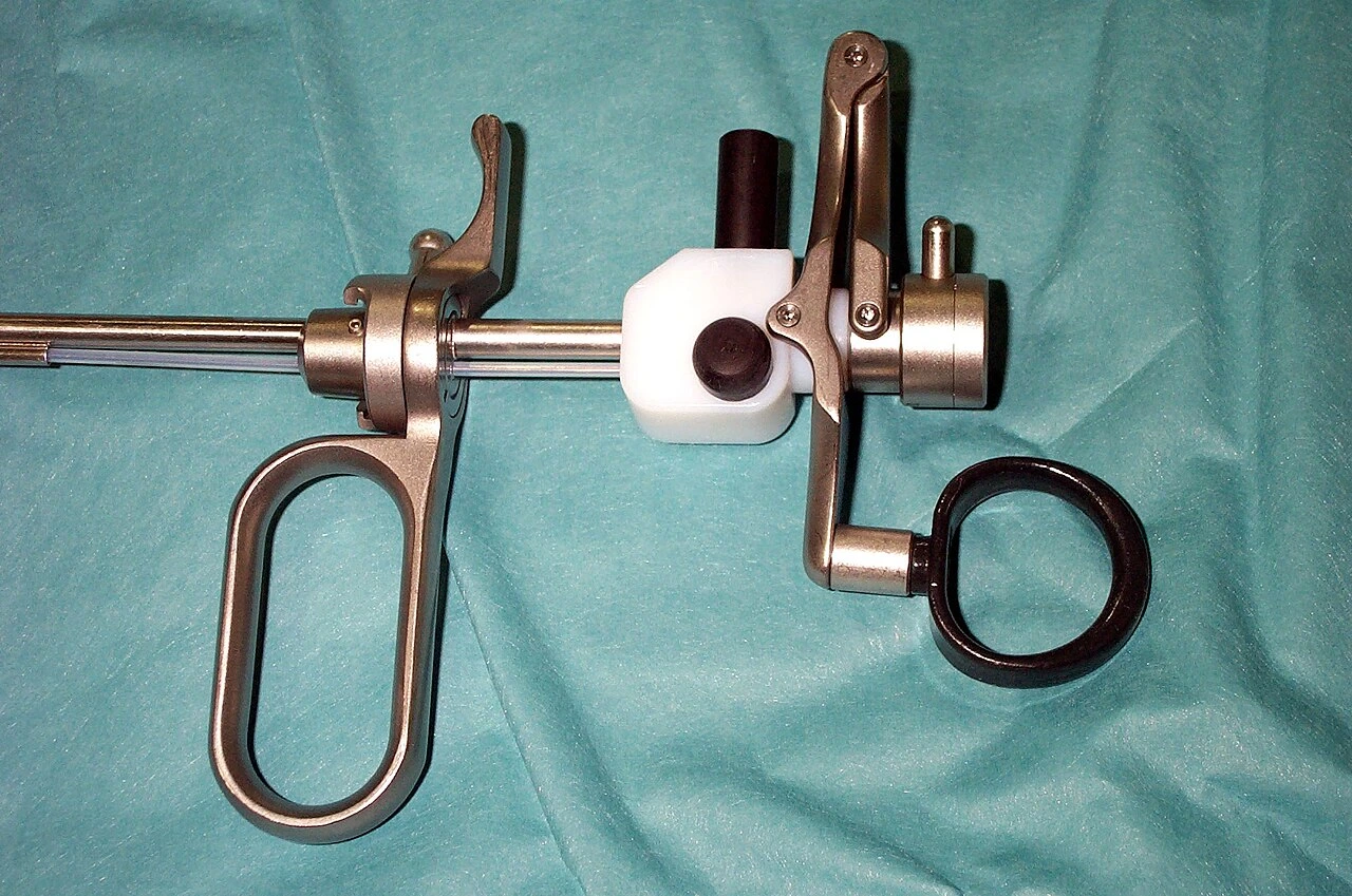 Two large finger rings flank a spring-and-lever assembly attached to a narrow rigid metal shaft.