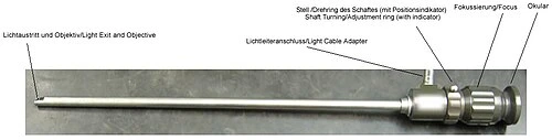 A slender straight metal viewing tube has a proximal eyepiece and a side connection, with component annotations in German.